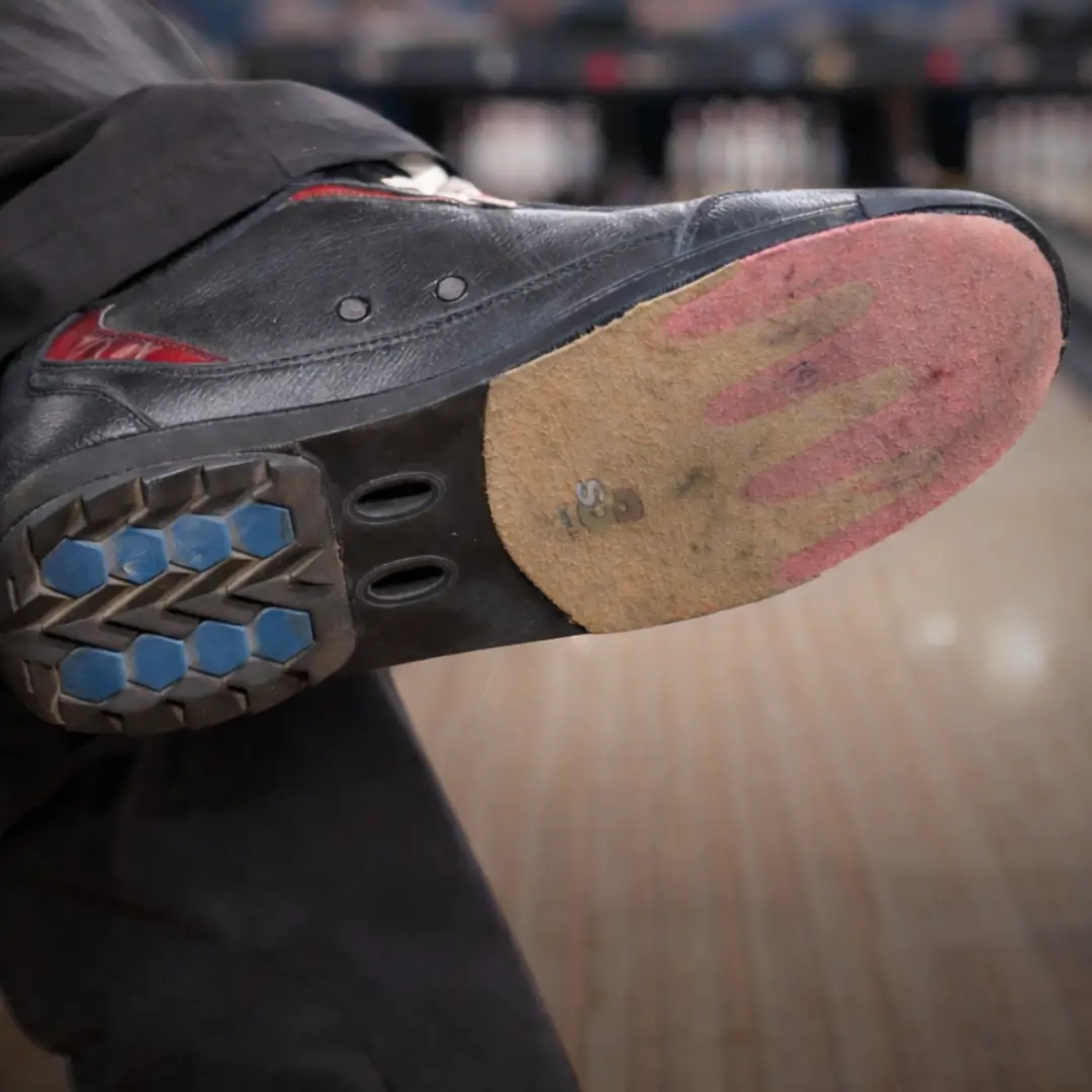 Close-up of a bowling shoe sole with interchangeable slide pad and heel on the approach, with a bowling ball blurred in the background.